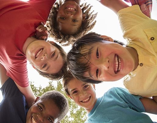Image looking up towards a group of diverse kids standing arm in arm in a huddle, smiling down at camera.