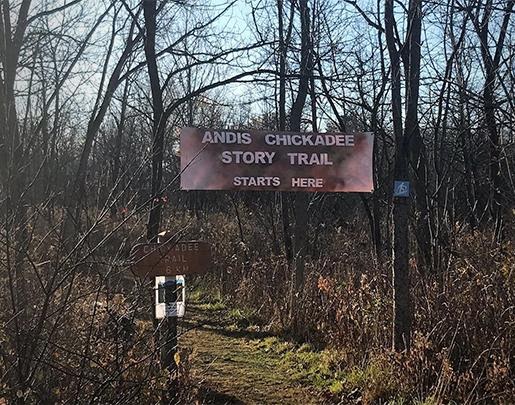 Large version of sign for Andis Chickadee Story Trail hanging above trail head with trees and nature and on either side of pathway.