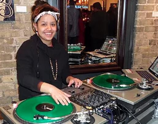 Large version of high-school age girl standing at DJ turn tables spinning a green record and smiling at camera